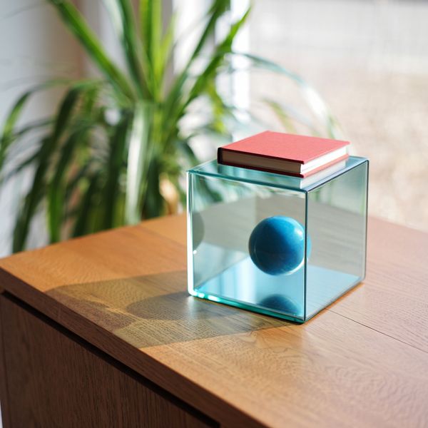 A glass cube on a wooden table. Inside the cube is a small blue sphere. On top of the cube sits a...