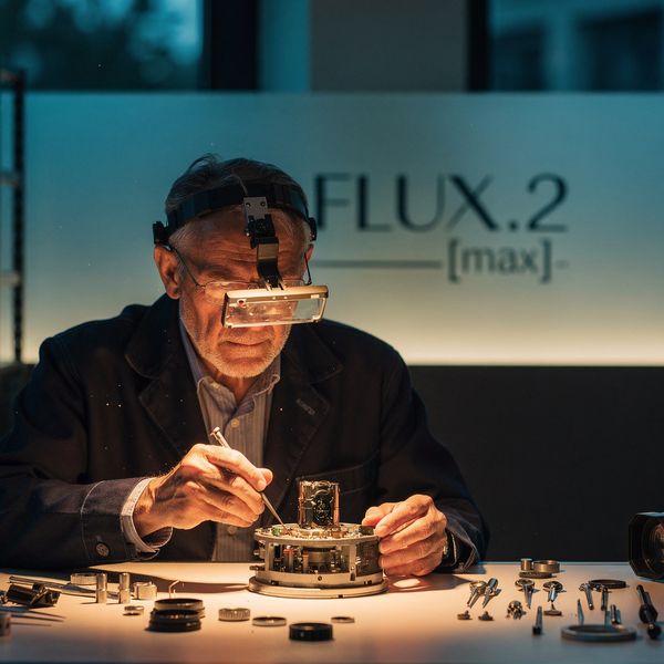 AI image generated by FLUX.2 [max] — prompt: A wide, cinematic shot of a sophisticated high-end camera lens repair shop at dusk. In the center, a master watchmaker-style craftsman works under a warm, focused spotlight on a complex optical assembly. In the blurred background, a sleek, minimalist frosted glass partition features the elegant, etched brand logo "FLUX.2 [max]" in professional typography. The scene is rendered with deep shadows, rich amber and teal color grading, and a shallow depth of field, emphasizing a hyper-realistic, polished aesthetic. 16:9 aspect ratio.