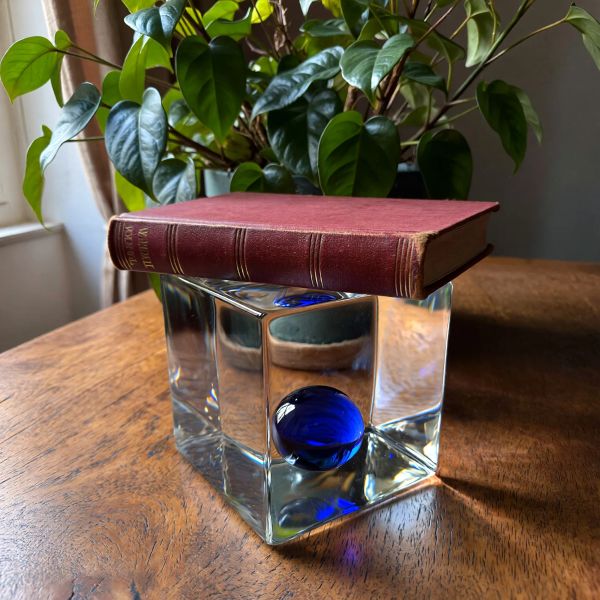 A glass cube on a wooden table. Inside the cube is a small blue sphere. On top of the cube sits a...