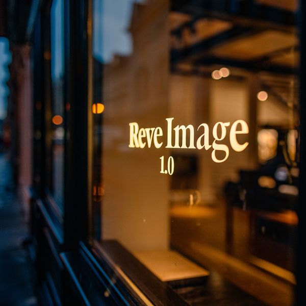 AI image generated by Reve Image 1.0 — prompt: Cinematic wide shot of a sophisticated, high-end boutique storefront at dusk. The warm, golden interior glow spills onto a rain-slicked cobblestone street. Centered in the large, polished glass window is an elegant, backlit gold leaf signage that reads "Reve Image 1.0" in a refined, modern serif typeface. Soft bokeh from city lights in the background, deep navy and amber color palette, professional architectural photography style, 16:9 aspect ratio.